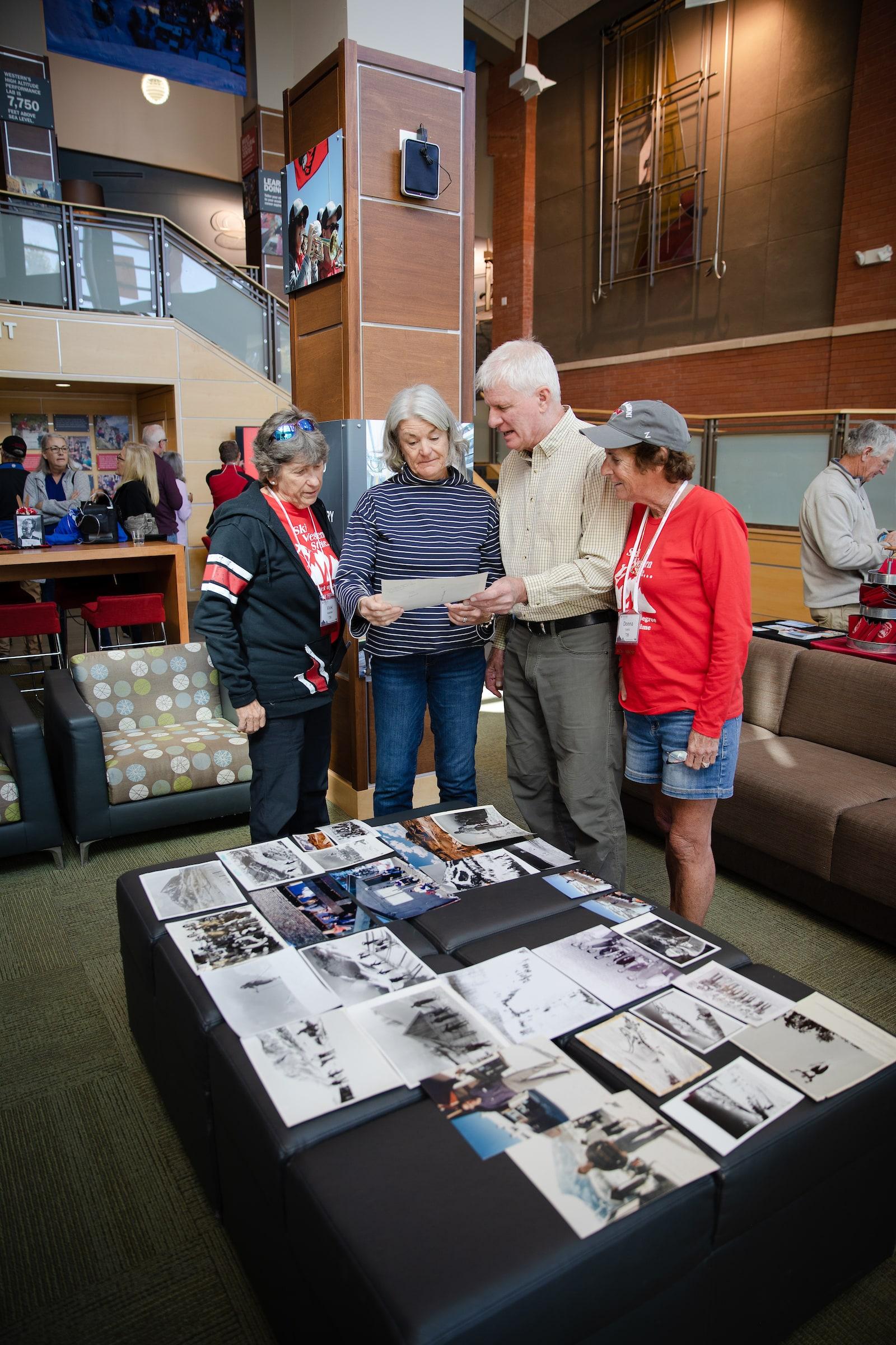 A group of four alumni look at a display of old photos on a table.